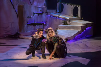 Puck in black jacket, pants and shoes decorated in flourescent swirls and with little goat horns in her head crouches next to a crouching Oberon, long blond hair, black and flourescent green cloak and pants, with the white rock set in the background